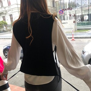 Op de afbeelding: Een zwart fluwelen vest met een wit shirt en een zwarte jeans. Het vest heeft twee lange banden die aan de voorkant kunnen worden vastgebonden.