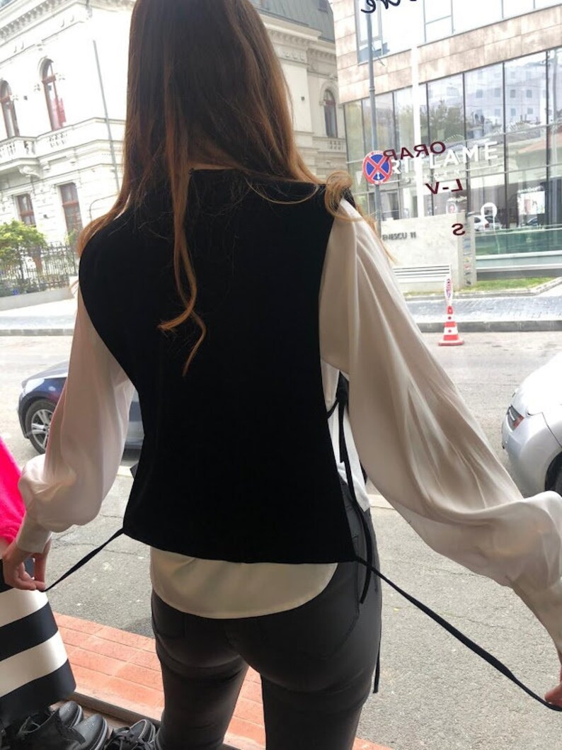 Op de afbeelding: Een zwart fluwelen vest met een wit shirt en een zwarte jeans. Het vest heeft twee lange banden die aan de voorkant kunnen worden vastgebonden.