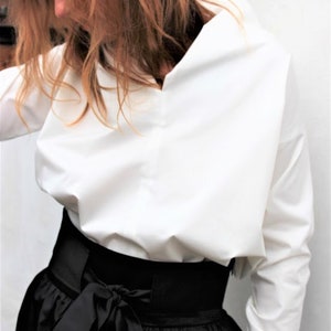 Può includere: Una blusa bianca a maniche lunghe con una vestibilità rilassata e un collo a V. La blusa è infilata in una gonna nera con una cintura larga e un fiocco.