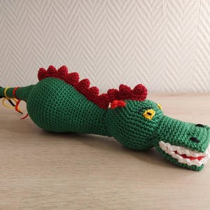 May include: A handmade green crochet dragon toy with red spines and yellow eyes. The dragon has a long tail with black strands and colorful accents. The mouth is white with red detailing.