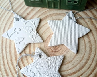 May include: Three white star-shaped ornaments with textured surfaces, each featuring a silver string loop. One star has a holly leaf pattern, one has vertical lines, and one has geometric motifs. A small green and white house sits behind them.