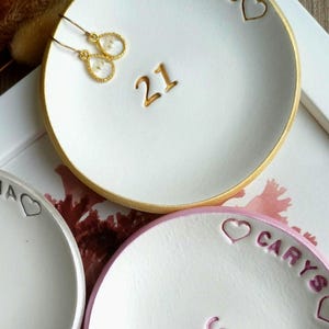 May include: Two round white ceramic trinket dishes with gold and pink painted rims. One dish features the name JESS, the number 21, and heart icons in gold. The other dish features the name CARYS, the number 16, and heart icons in pink.