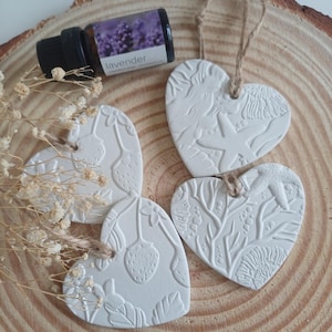 May include: White heart-shaped ornaments with embossed designs of strawberries, starfish, and coral, suspended by twine. A small bottle of lavender essential oil is placed nearby. The ornaments are on a wooden slice.