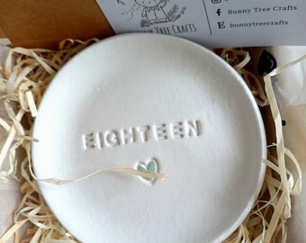 May include: A white ceramic dish with the word "EIGHTEEN" embossed on its surface, accompanied by a small green heart. The dish is nestled in a bed of straw inside a cardboard box. A card with a bunny illustration and the text "Bunny Tree Crafts" is visible.
