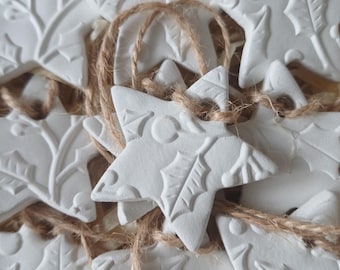 May include: A collection of white star-shaped ornaments made of clay, each featuring an embossed holly leaf and berry pattern. Every star has a hole at the top threaded with a piece of natural brown jute twine for hanging purposes.