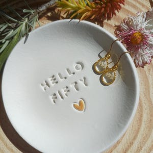 May include: A white ceramic trinket dish with the words "HELLO FIFTY" embossed on the surface, along with a gold heart. Two gold earrings with clear teardrop-shaped pendants containing small white flowers rest on the dish.