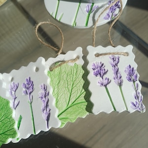 May include: Three rectangular white clay ornaments with scalloped edges, each featuring embossed lavender sprigs in purple and green, or a large green leaf imprint. Each ornament includes a brown twine loop for hanging on the top edge.