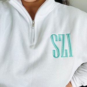 May include: White quarter-zip sweatshirt with a silver zipper. The sweatshirt features the embroidered initials "SZI" in a teal color on the front. The sleeves are long and the material appears to be a soft cotton blend.