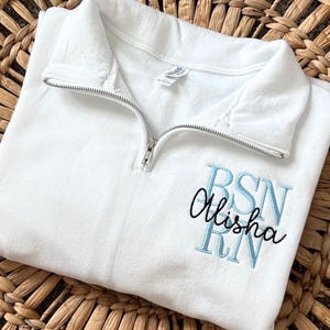 May include: White quarter-zip sweatshirt with a silver zipper. The sweatshirt has the embroidered text "BSN RN" in light blue, with the name "Alisha" in cursive below. The sweatshirt is on a woven, brown surface.