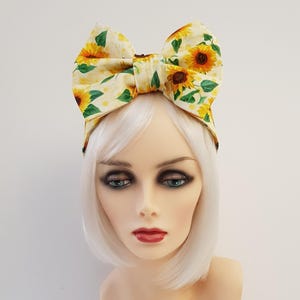 May include: A fabric headband featuring a large, structured bow on top. The material is a light cream colour with a repeating pattern of yellow sunflowers, brown centres, and green leaves, accented by small, faint yellow polka dots throughout the design.