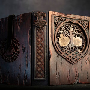 Puede incluir: Una caja de madera con una correa de cuero y tallas intrincadas. El panel frontal presenta un diseño de árbol de la vida con un fondo dorado. El panel lateral tiene un símbolo nórdico y un diseño de martillo.