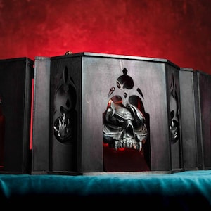 Può includere: Una scatola di legno nera con un teschio e due figure all'interno. La scatola è aperta e il teschio è illuminato da una luce rossa.