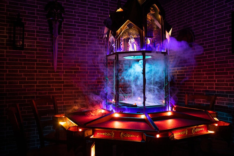 May include: A dark wooden table with a red felt top and a decorative, ornate, multi-level, wooden structure in the center. The structure is lit with blue and purple lights and has a glass dome with smoke inside. The table is surrounded by four chairs.