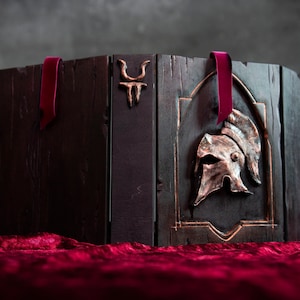 Puede incluir: Una caja de madera oscura con un diseño de casco de metal en la parte delantera. La caja tiene cintas de terciopelo burdeos atadas a los lados.