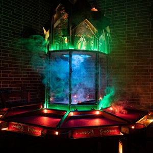 May include: A dark wooden game table with a red felt surface and a green and blue stained glass tower in the center. The tower is surrounded by smoke and lights.