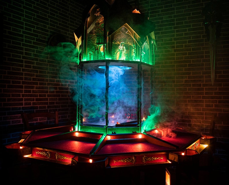 May include: A dark wooden game table with a red felt surface and a green and blue stained glass tower in the center. The tower is surrounded by smoke and lights.