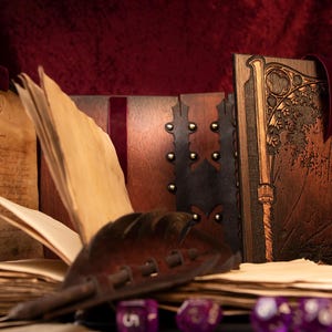 May include: A collection of handcrafted, antique-style journals and books with aged paper pages. The journals feature intricate wooden covers with metal studs and decorative carvings. A leather quill and purple dice are in the foreground.