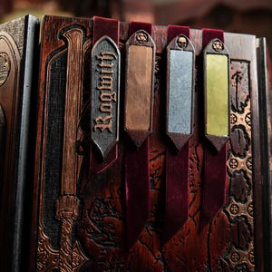 Könnte beinhalten: Nahaufnahme eines Holzbuchs mit kunstvollen Schnitzereien und dekorativen Metall-Lesezeichen. Die Lesezeichen sind in verschiedenen Farben gehalten, darunter Kupfer, Silber und Gold, und sind an roten Samtbändern befestigt. Der Buchrücken trägt die Aufschrift "Legends".
