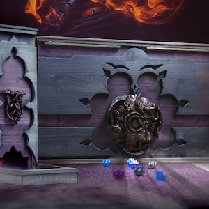 May include: A wooden dice box with a purple interior and grey exterior. The box features ornate carvings and a bronze-coloured emblem. Several dice are scattered in front of the box, and a lit candle is to the right.