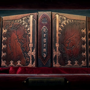 May include: A wooden book-style box with the word "Legend" on the spine. The box is brown with ornate gold-colored accents and a map-like design. It rests on a red velvet surface.
