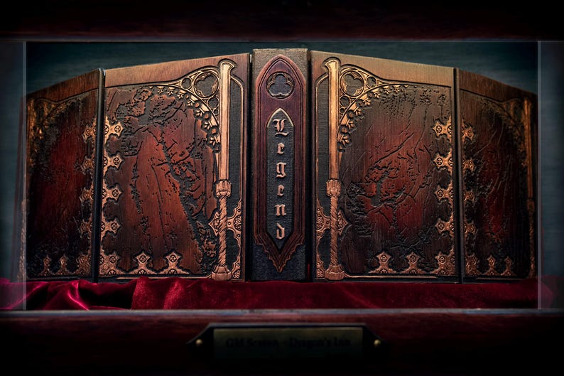 May include: A wooden book-style box with the word "Legend" on the spine. The box is brown with ornate gold-colored accents and a map-like design. It rests on a red velvet surface.