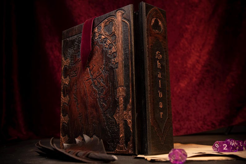 May include: A large, ornate wooden book with intricate carvings and a burgundy ribbon bookmark. The book's cover features detailed relief work and a metallic gold accent. The spine reads "Skalbor" in a gothic font. A feather and dice are in the foreground.