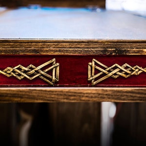 May include: Close-up of a wooden table top with a red velvet inlay. The inlay features two gold metal Celtic knot designs.