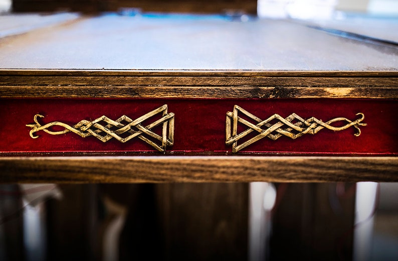 May include: Close-up of a wooden table top with a red velvet inlay. The inlay features two gold metal Celtic knot designs.