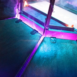May include: A close-up of a wooden box with a hinged lid. The box is lit with purple and blue lights, creating a dramatic effect.