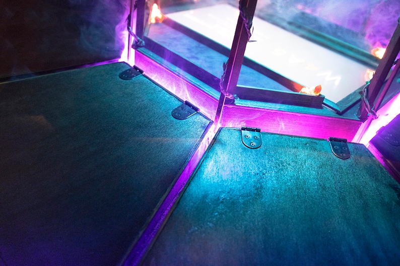 May include: A close-up of a wooden box with a hinged lid. The box is lit with purple and blue lights, creating a dramatic effect.