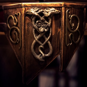May include: Close-up of a wooden table corner with intricate gold metalwork. The design features two intertwined snakes with their heads facing each other.