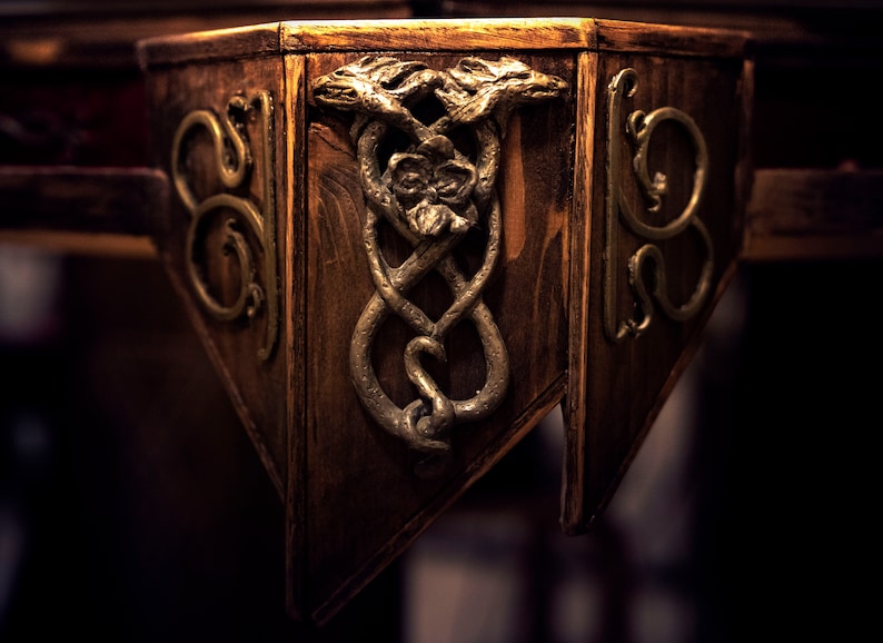 May include: Close-up of a wooden table corner with intricate gold metalwork. The design features two intertwined snakes with their heads facing each other.