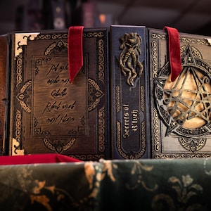 May include: A set of three leather-bound books with ornate gold detailing and red ribbon bookmarks. The books are titled "Secrets of the Witch" and "The Book of Shadows".
