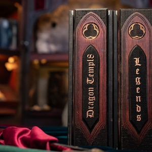 Puede incluir: Dos libros de tapa dura con cubiertas marrones oscuras y detalles burdeos. El libro de la izquierda dice "Dragon Temples" en letras doradas, y el libro de la derecha "Legends". Los libros se exhiben sobre una superficie oscura con una tela roja en primer plano.