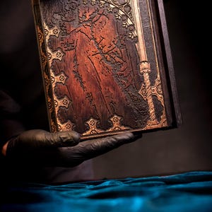 May include: A dark wooden book with intricate carvings and gold-colored accents. The cover features a detailed relief design of a figure and a sword. The book is held against a dark background, highlighting the detailed craftsmanship.