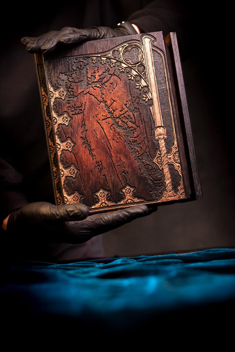 May include: A dark wooden book with intricate carvings and gold-colored accents. The cover features a detailed relief design of a figure and a sword. The book is held against a dark background, highlighting the detailed craftsmanship.