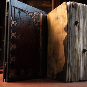 May include: An open, antique-style book with dark brown wooden covers and ornate copper-coloured detailing. The aged pages have a yellowed, stained appearance, and the book is secured with triangular metal clasps. The book is likely a prop or decorative item.