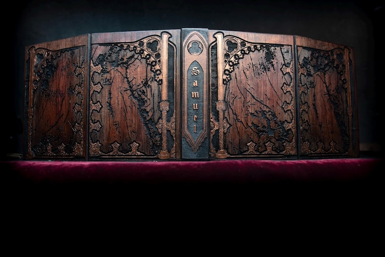 May include: A wooden book-shaped box with intricate carvings and the name "Samuel" on the spine. The box is brown with gold accents and rests on a red velvet surface. The design features gothic-style details.
