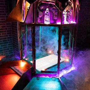 May include: A wooden structure with a glass enclosure, illuminated with purple and blue lights. The structure has a small, white, rectangular screen inside the glass enclosure. The structure is surrounded by smoke.
