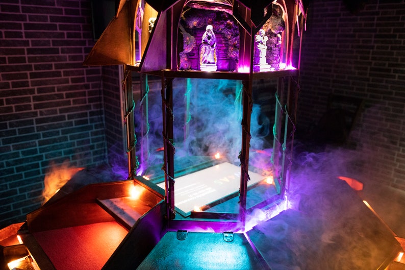 May include: A wooden structure with a glass enclosure, illuminated with purple and blue lights. The structure has a small, white, rectangular screen inside the glass enclosure. The structure is surrounded by smoke.