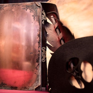 May include: Close-up of a vintage book with a copper-colored cover and ornate metal accents. The book's cover has a distressed appearance, with visible wear and tear. The binding is dark wood with decorative metal studs.