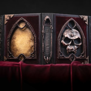 Puede incluir: Un libro de madera oscura con un diseño de calavera y un rollo. El libro está cerrado y tiene un forro de terciopelo rojo. El libro está sobre una superficie de terciopelo rojo.