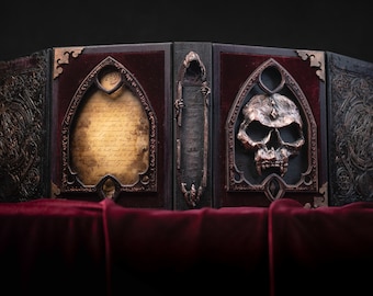 May include: A dark wooden book with a skull and a scroll design. The book is closed and has a red velvet lining. The book is on a red velvet surface.