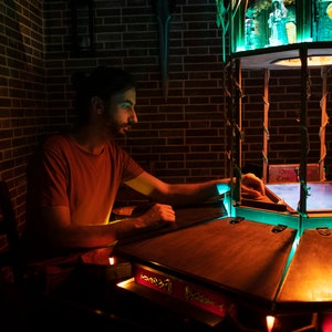 May include: A person sits at a wooden table with a glowing, multi-sided game board. The board has a blue, glowing surface and is surrounded by a wooden frame. The game board is lit by a green light from above.