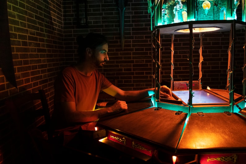 May include: A person sits at a wooden table with a glowing, multi-sided game board. The board has a blue, glowing surface and is surrounded by a wooden frame. The game board is lit by a green light from above.