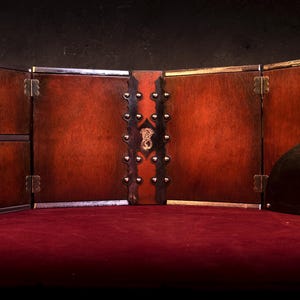May include: A wooden tabletop game screen with a dark brown finish. The screen is divided into panels, with brass hinges and decorative studs. A semi-circular panel with a cut-out design is on the right. The screen is set on a red surface.