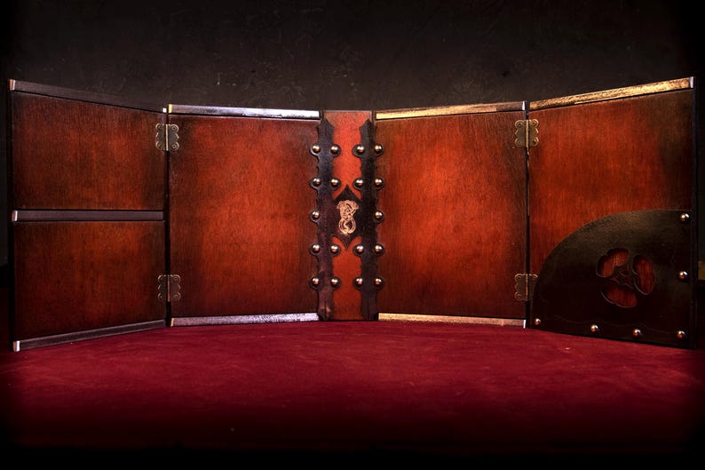 May include: A wooden tabletop game screen with a dark brown finish. The screen is divided into panels, with brass hinges and decorative studs. A semi-circular panel with a cut-out design is on the right. The screen is set on a red surface.
