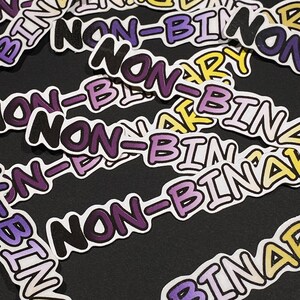 Peut inclure: Plusieurs autocollants avec l'inscription "NON-BINARY" dans une police stylisée. Le texte est de différentes couleurs, notamment violet, jaune et blanc, avec un contour noir. Les autocollants sont disposés sur une surface sombre.