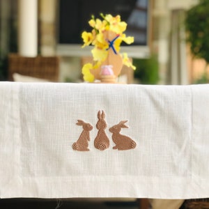 Può includere: Un runner da tavolo in lino bianco con coniglietti marroni ricamati. I coniglietti sono in fila, rivolti in avanti.