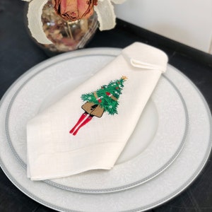 May include: White linen napkin with a red and green embroidered design of a person carrying a Christmas tree.